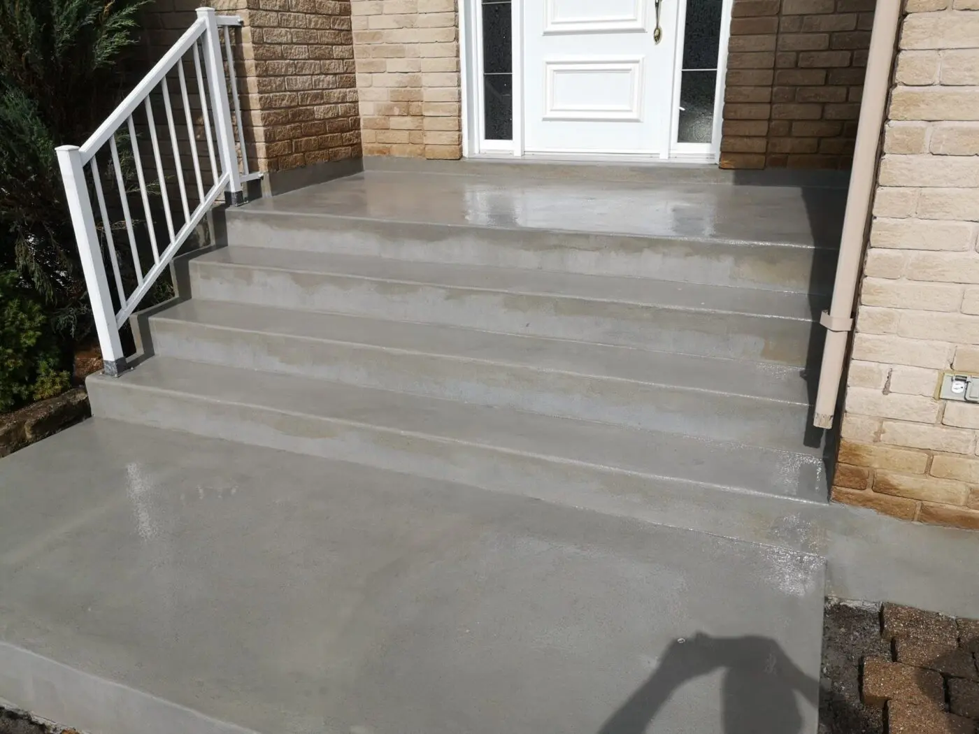A set of wide concrete steps, crafted by an affordable concrete contractor, leads up to a white-paneled front door of the brick house. The steps have a smooth finish and are flanked by white railings on the left side. A shadow of a person taking the photo is visible in the foreground.