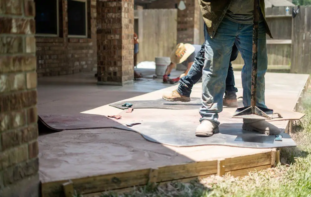 Two contractors lay concrete in the backyard of a brick house. One worker flattens the surface with a tool, while the other is on their knees, working on the edge. The outdoor space is partly shaded, with tools and materials visible around them, showcasing a home improvement project in progress.