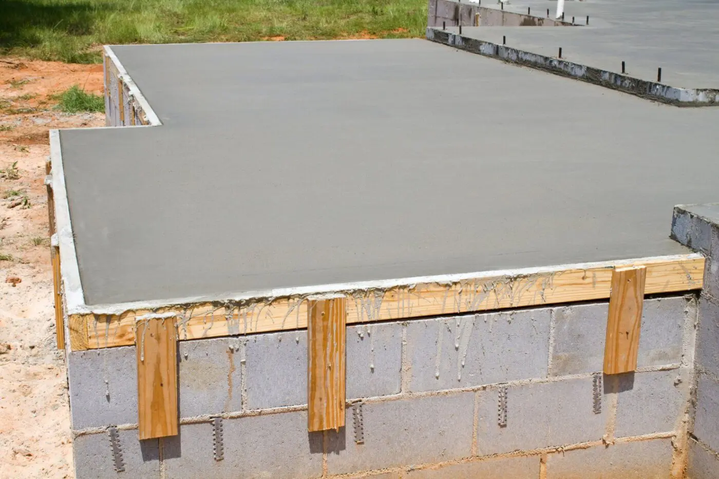 A close-up view of a construction site in Broward County shows a recently poured and smooth concrete foundation. The concrete, ideal for driveways, is laid over a base of cinder blocks, reinforced with wooden planks on the sides. The ground around the site is a mix of dirt and grass.
