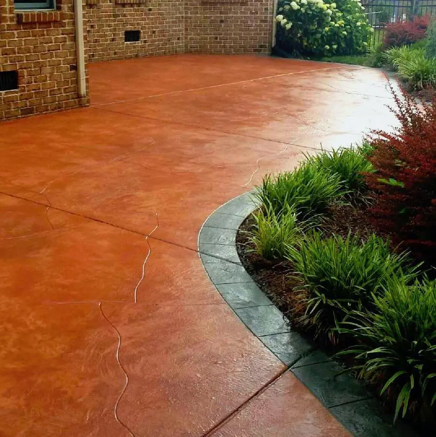 A patio with a reddish-brown stamped concrete surface, bordered by a darker, slightly curved edge. Adjacent to the patio, there are landscaped bushes and small plants, with the corner of a red-brick building visible in the background—an exceptional job by an affordable concrete contractor from Broward County.