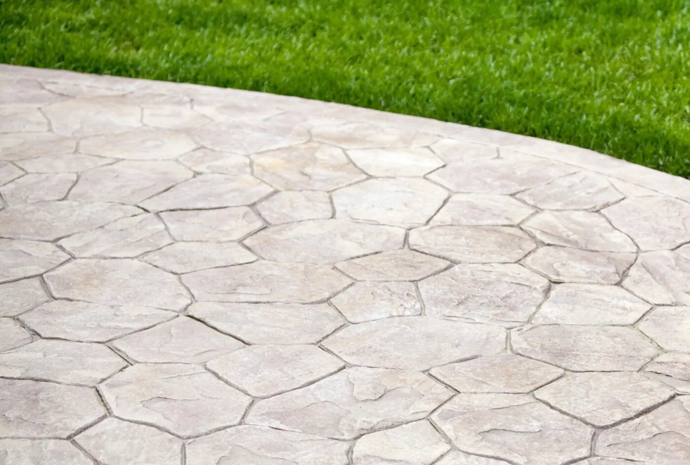 A close-up of a decorative concrete walkway featuring irregularly shaped stones in a light color. The walkway curves to the right and is bordered by a neatly trimmed, lush green lawn, showcasing the craftsmanship of skilled contractors.