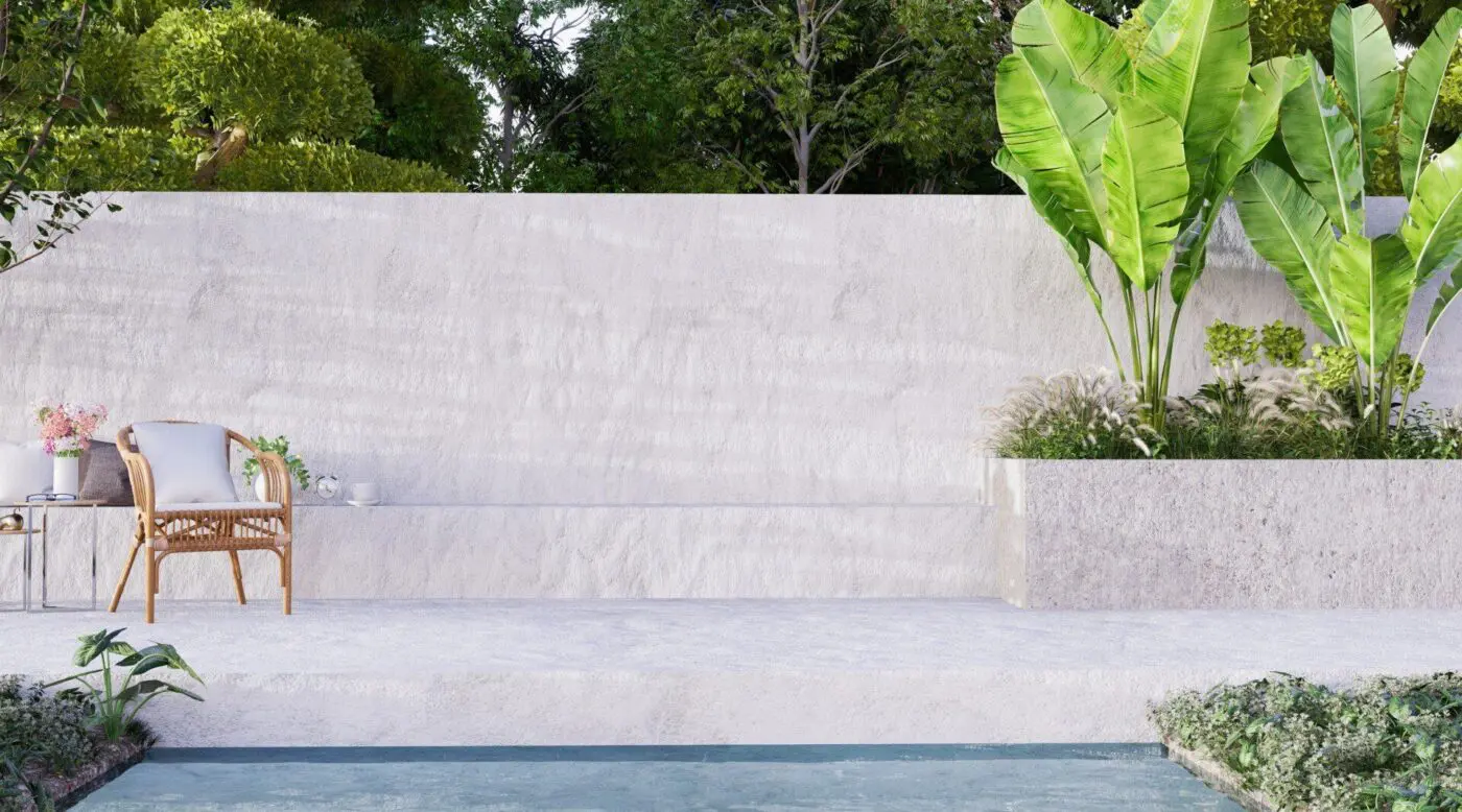 A minimalist outdoor patio with a wicker chair and a small side table holding flowers and a cup. There is a white stone wall in the background, featuring Lauderdale Concrete from Miramar FL, with lush green plants and trees, and a small blue pool in the foreground.