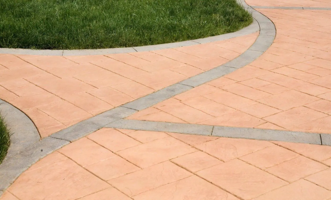 A paved pathway with a light peach-colored tile pattern and gray border, intersecting and curving through a well-maintained grassy area in Pompano Beach, FL. The curved lines create a geometric design on the ground, offering cost-effective concrete solutions for both residential and commercial properties.
