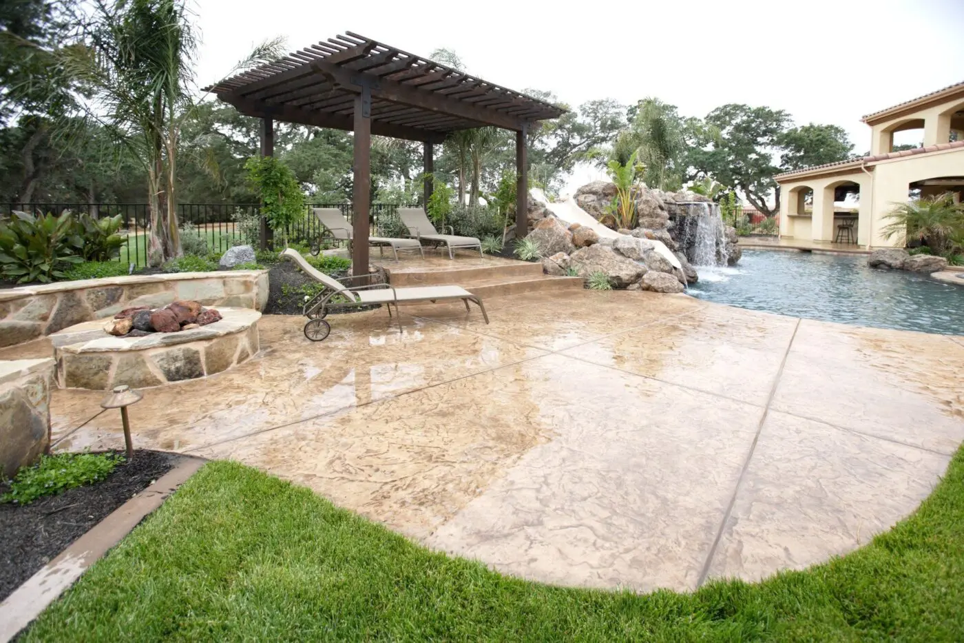 A luxurious backyard features a stamped concrete patio by Lauderdale Concrete with a poolside pergola, several lounge chairs, and lush landscaping. A stone fire pit is positioned on the left, and a rock waterfall cascades into the pool. The area is surrounded by greenery and trees. Contact Lauderdale Concrete for a free quote today!