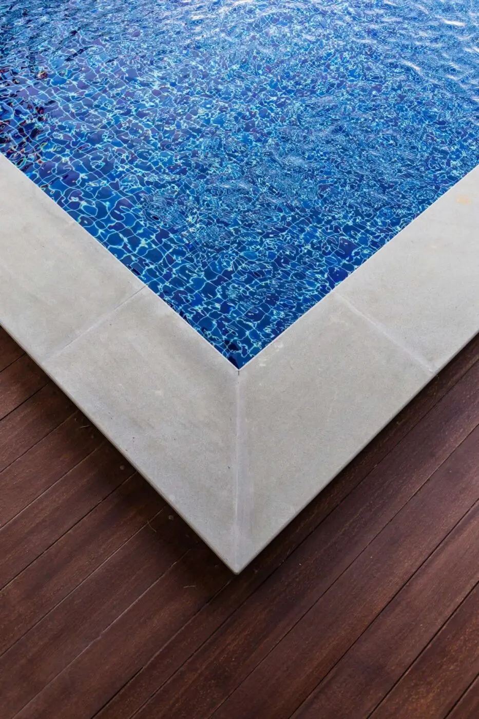 Close-up of a swimming pool corner with vibrant blue water mosaic tiles. The pool's edge is bordered by light-colored stones, adjacent to a wooden deck with a rich brown hue, highlighting the contrast between the deck and the pool. Expertly designed by Lauderdale Concrete in Coral Springs FL.