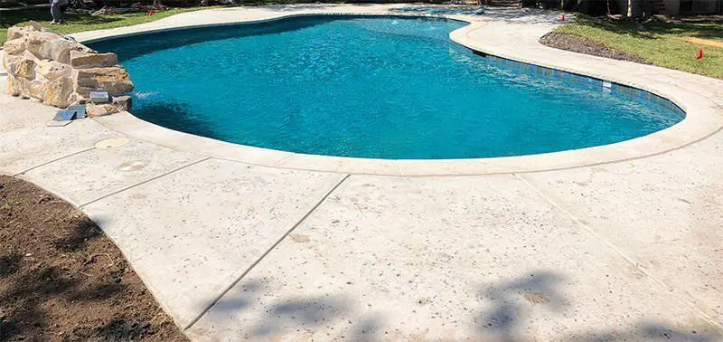 A clear blue swimming pool with a curvy shape is surrounded by decorative concrete pavement. To the left, a small rock feature is placed near the edge of the pool. The surroundings include grass and some shadows, suggesting a sunny day.