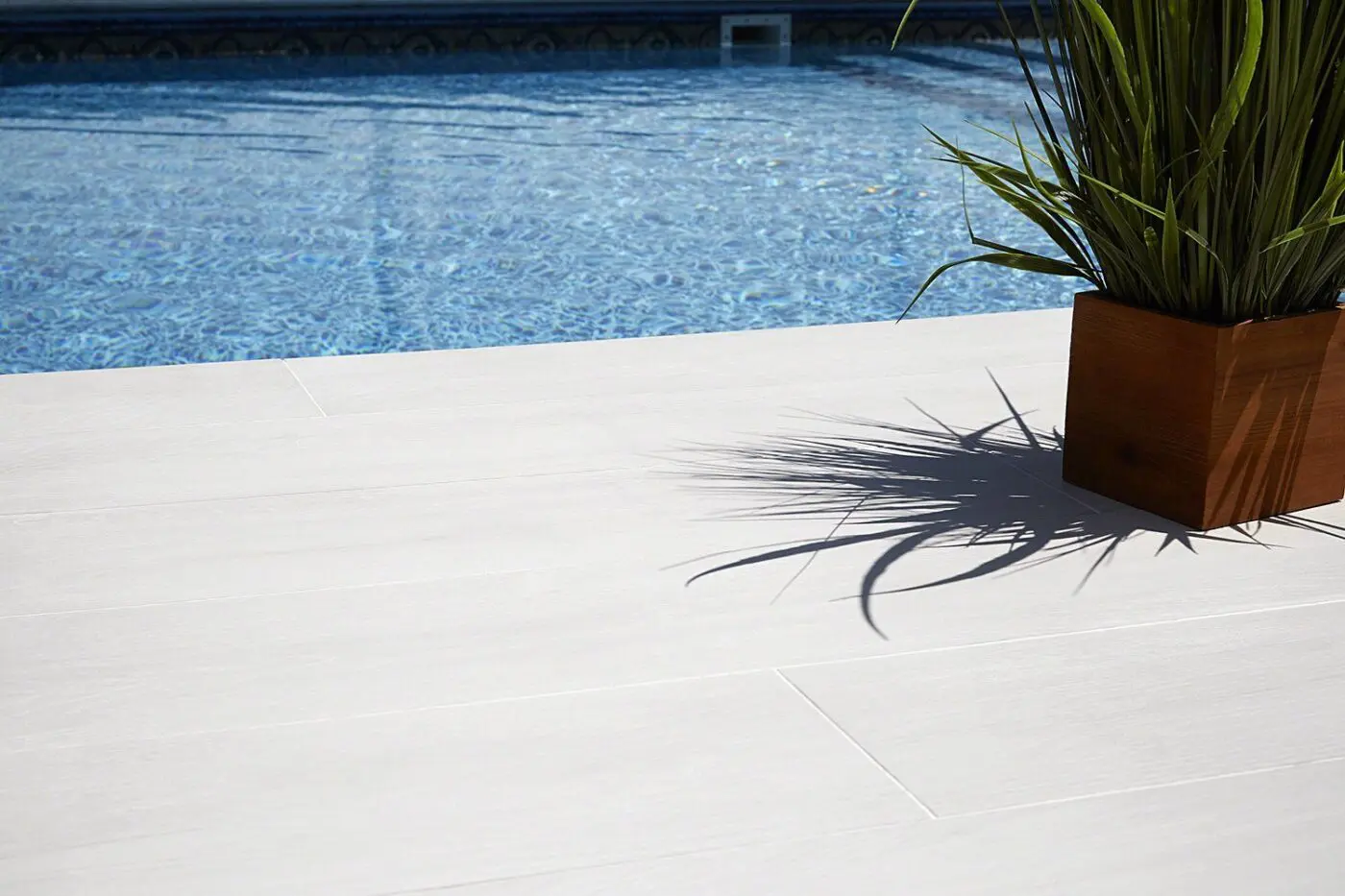 Close-up of a poolside area in Pembroke Pines, FL, with light-colored tile flooring. A potted green plant in a wooden planter casts a shadow on the tiles, and the clear blue water of the pool is visible in the background. Contact us for a free quote to enhance your space with our concrete solutions.