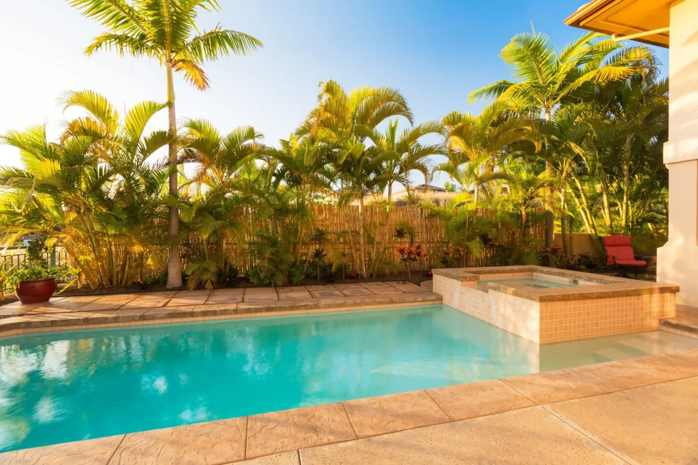 A serene backyard pool area with crystal clear water, surrounded by tall palm trees and lush greenery in Weston, FL. Adjacent to the pool is a raised hot tub. The setting is tranquil under a clear, blue sky with a warm and inviting atmosphere. Contact us for concrete services or a free quote today!