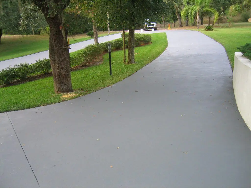 A smooth, curved concrete driveway, expertly crafted by Lauderdale Concrete contractors, runs through a grassy area with trees and shrubs on both sides. A small vehicle is visible in the distance near the end of the driveway. The scene is set in a well-maintained, green landscape typical of Coral Springs FL.