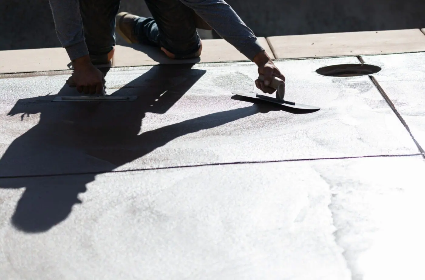 A person is smoothing wet concrete with a trowel on a sunny day in Davie, FL. Their shadow is visible on the surface, highlighting their precise movements. Wearing a long-sleeved shirt, their hands grip the trowel handles expertly, showcasing Lauderdale Concrete Contractors' decorative concrete services.