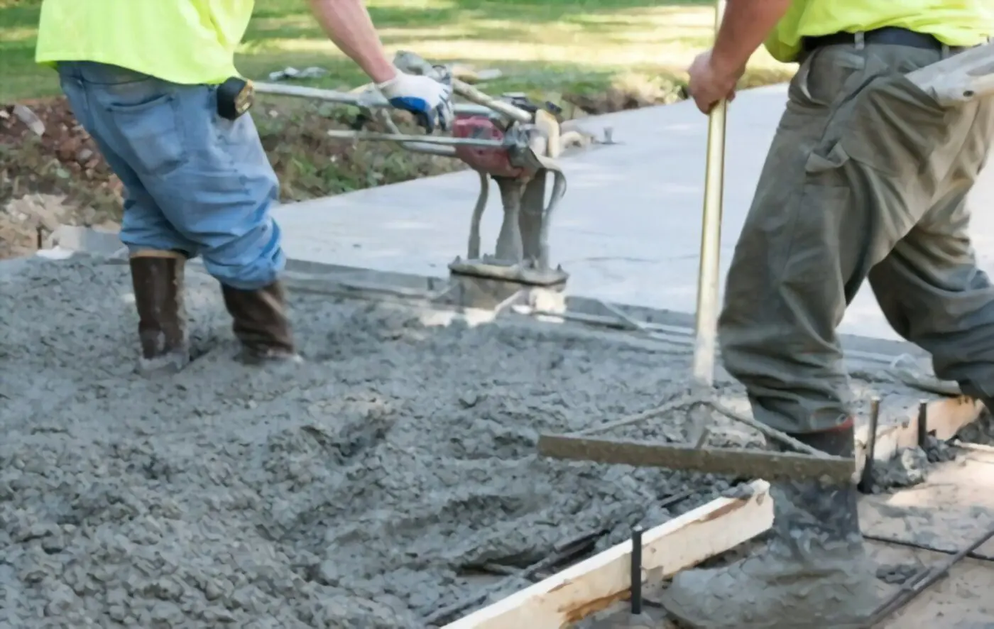 Two construction workers in protective gear, including gloves and boots, are working with tools to smooth and level wet concrete on a sidewalk. The scene is outdoors, with visible grass and trees in the background, as part of a larger Lauderdale Concrete project in Miramar FL.