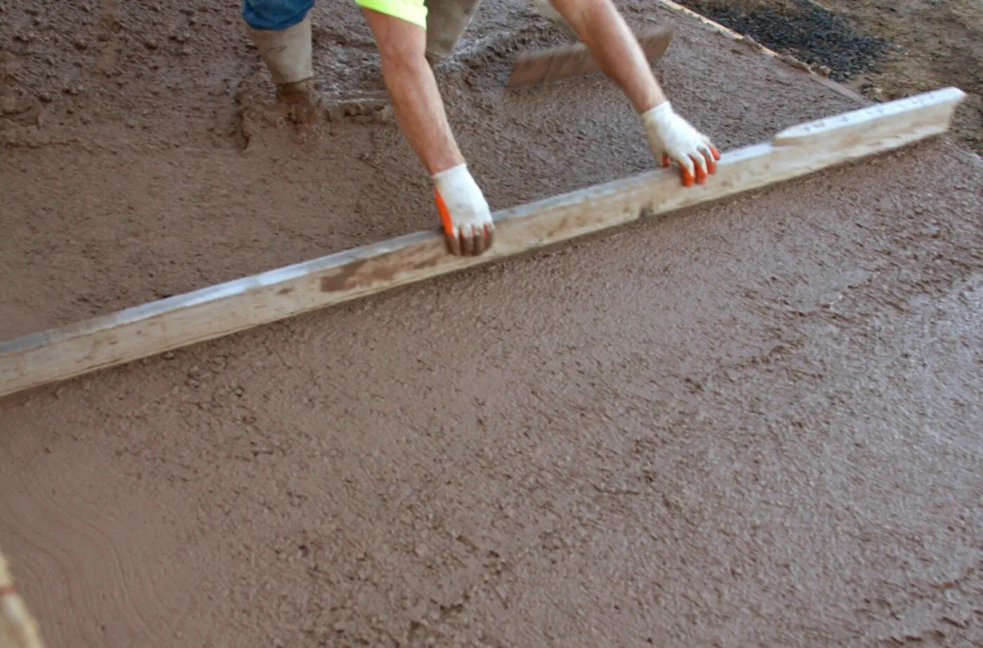 A person wearing a neon green shirt and white gloves spreads wet concrete with a long wooden board at a construction site in Pompano Beach, FL. Their lower half is visible, with legs in boots standing in the concrete. The freshly poured surface is being smoothed, ensuring cost-effective concrete solutions.