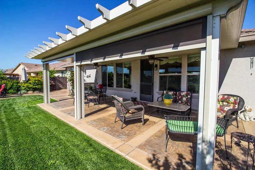 A covered patio features outdoor furniture, including a sofa with floral cushions, a glass-top table, and various chairs. This inviting outdoor space is shaded by a pergola and surrounded by a well-manicured lawn. The house and clear blue sky are visible in the background, completing the serene setting.