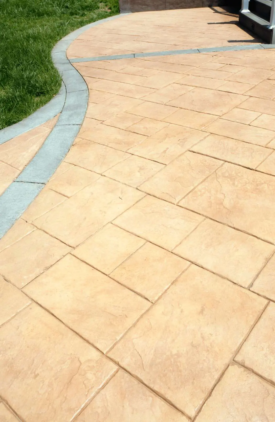 A patio area with large, rectangular, beige tiles arranged in a staggered pattern. A curved, narrow gray strip separates the concrete patio from a grassy area in the upper left corner. Stairs leading to a higher platform are visible in the background, showcasing the work of resurfacing professionals.