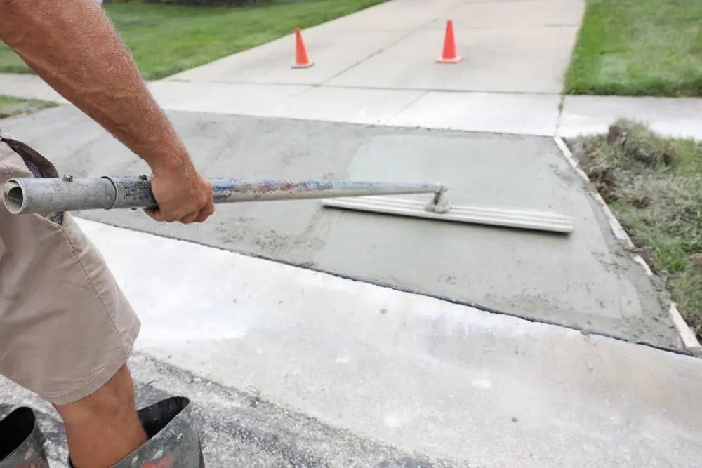 A person is smoothing out a freshly laid concrete sidewalk with a long-handled trowel. There are two red traffic cones placed on the paved road nearby, and the surrounding area is green grass. For quality concrete driveways, consult your local concrete driveway contractor and get free estimates today.