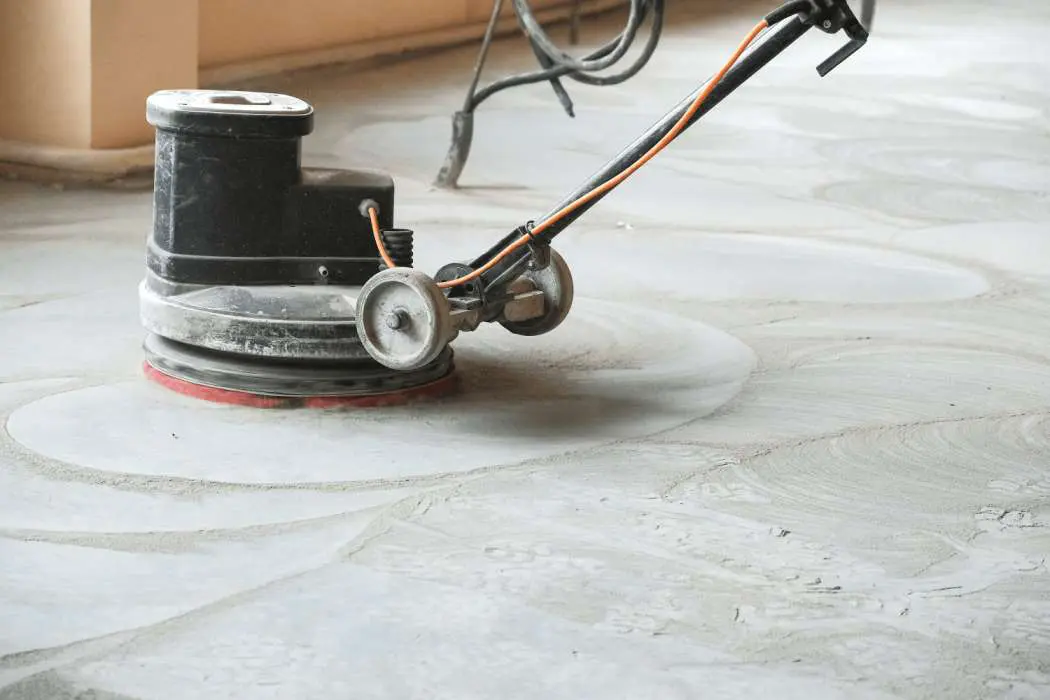 A floor buffer machine is in use on a cement floor during a large-scale concrete job. The circular scrubbing pad is rotating, and the machine's cord is visible. The floor appears to have recently been polished, showing wet and dry patches.