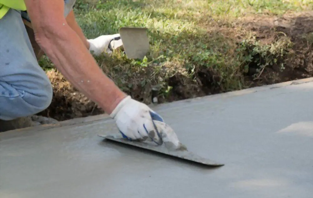 A concrete driveway contractor, wearing gloves, smooths wet concrete with a trowel on a grassy area. The individual's arm is extended, and there is a small shovel resting on the grass nearby, ensuring quality concrete driveways for all clients.