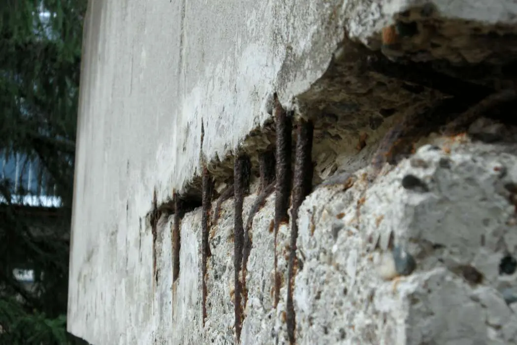 A close-up of a deteriorating concrete structure shows exposed, rusted rebar. The weathered and cracked surface is missing pieces, revealing corroded metal reinforcement bars underneath. A blurred tree is visible in the background, hinting at the need for commercial concrete services to restore integrity.