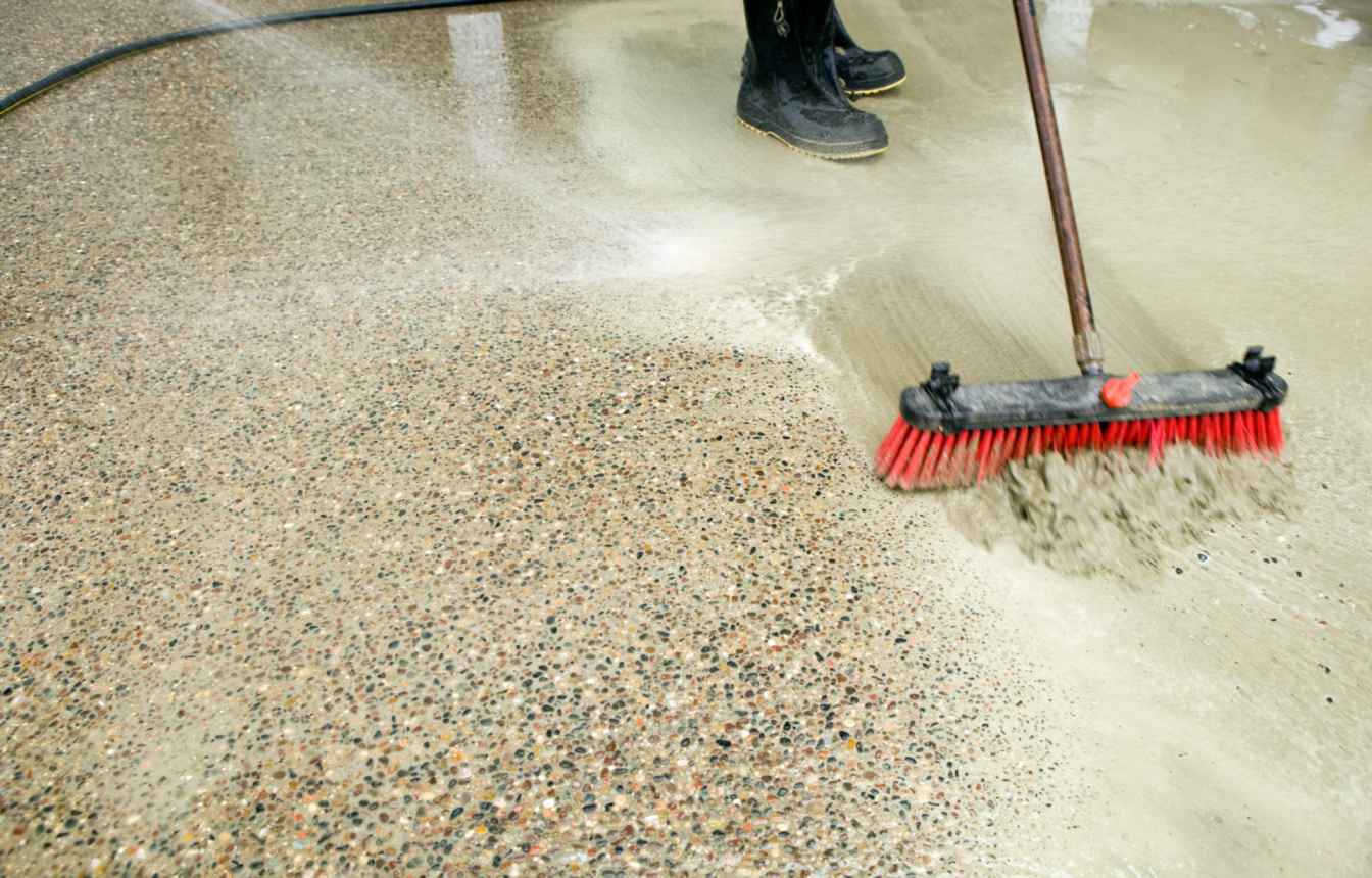 A resurfacing professional wearing black boots sweeps a wet, gravelly floor with a long-handled broom with red bristles.