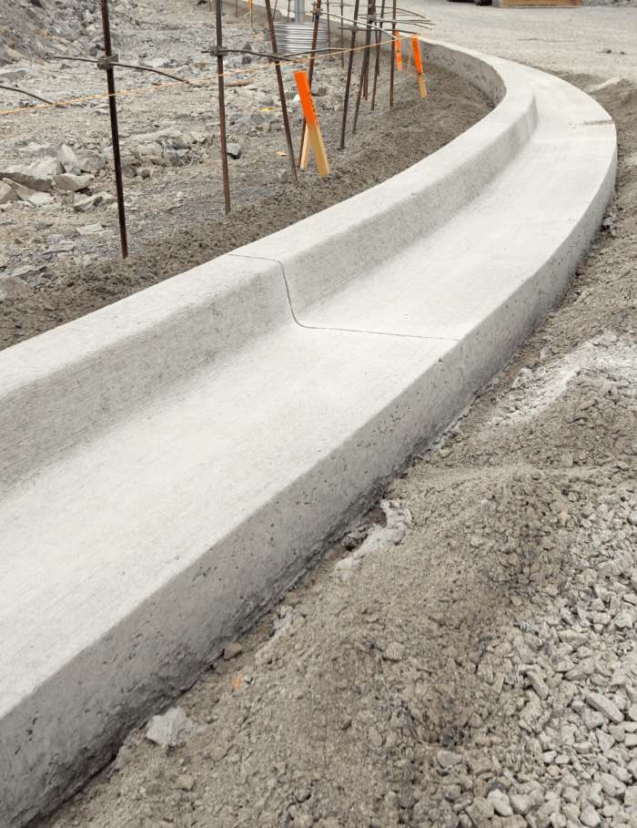 A freshly poured concrete curb in a construction area, with orange construction markers in the background. The curb curves gently and is surrounded by rough, gravelly ground. Scaffolding is also visible in the upper part of the image, thanks to the reliable concrete contractors known for their exceptional work in Broward County.
