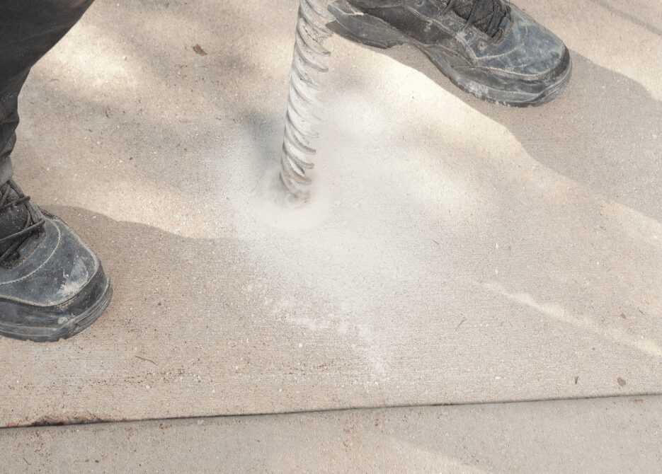 A person wearing work boots is using a large drill bit to make a hole in a concrete surface, likely as part of slab lifting or concrete restoration. Dust surrounds the drilling area, indicating active work. Shadows from the person add depth to the image, highlighting the outdoor setting in Broward County.